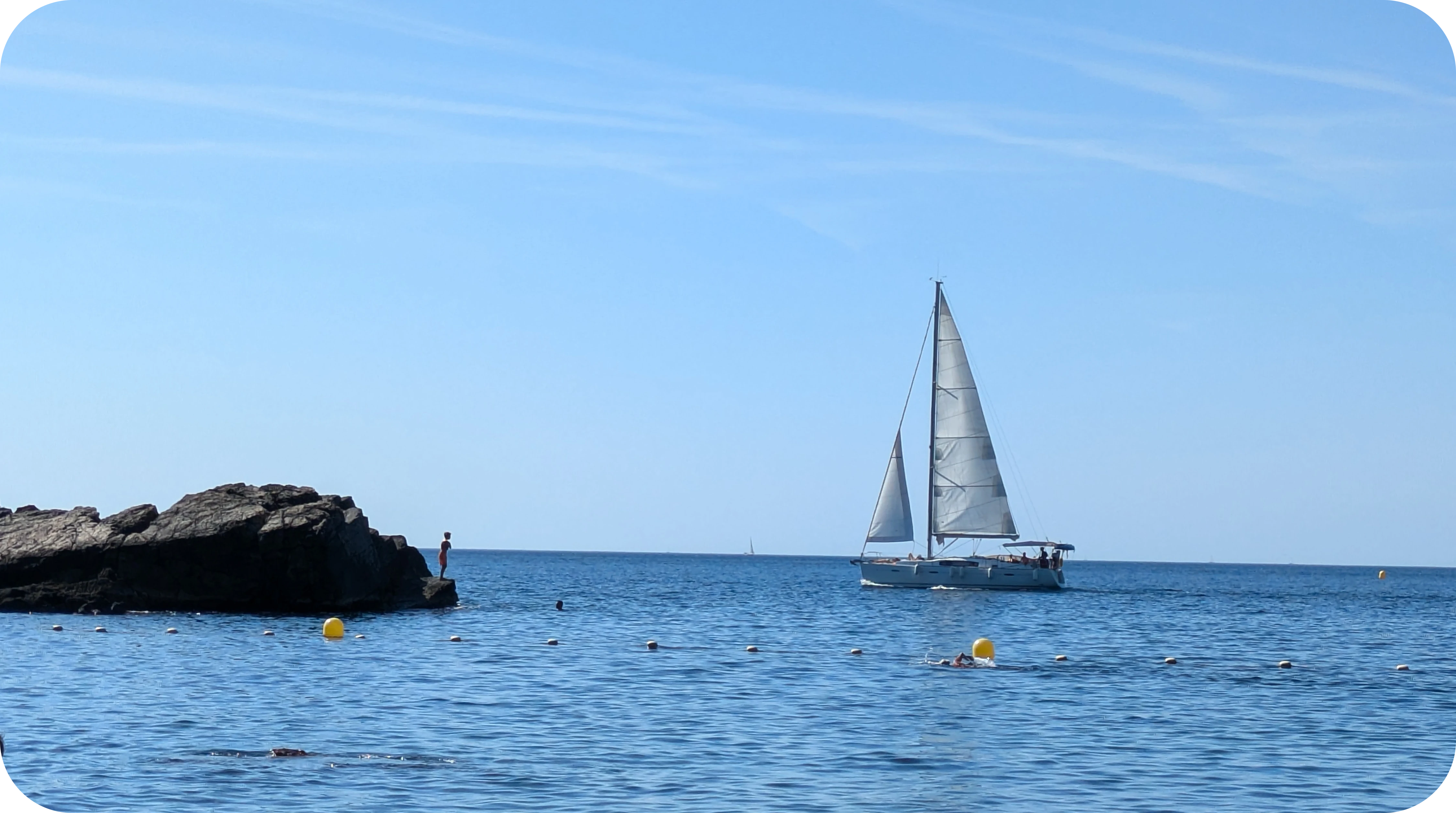 Une photo de la ligne d´horizon sur la mer, on y voit un voilier passer derrière un rocher.